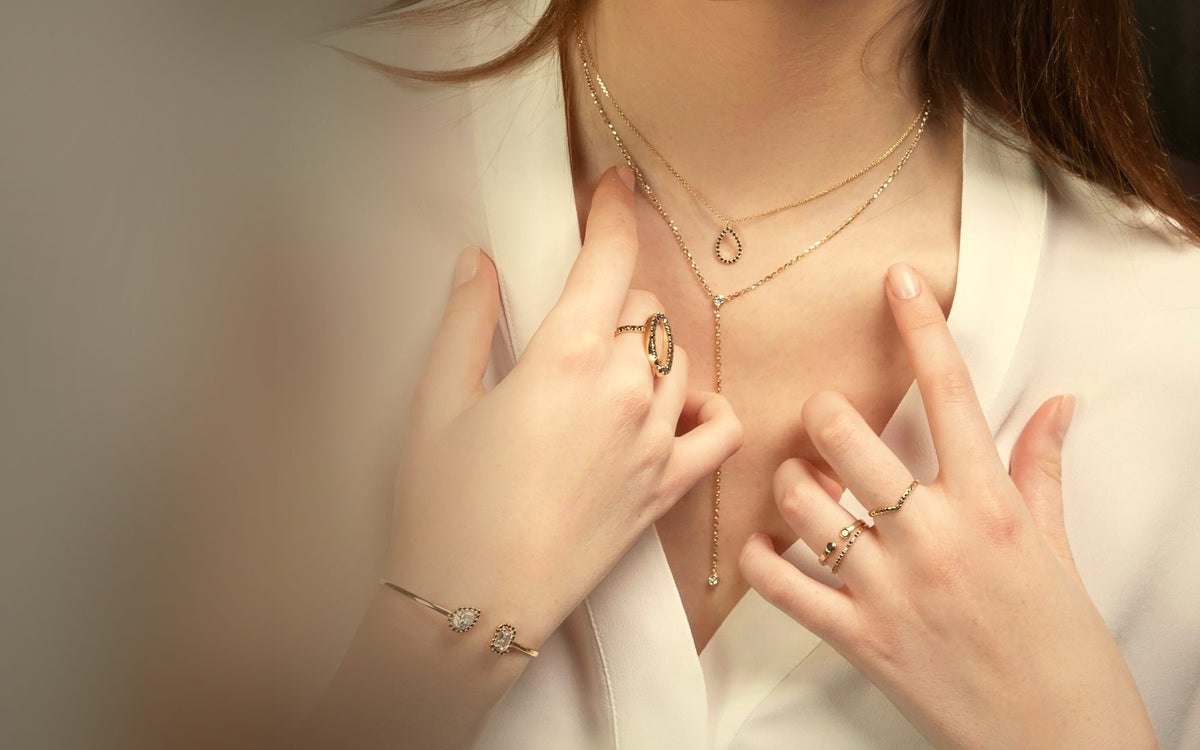Hands at an open white collar wearing two fine gold necklaces, stacked gold rings and an open gold bangle, in warm light
